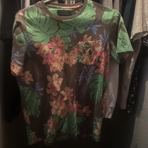 Blommig t-shirt från Polo - Snygg t-shirt från Polo med ett färgglatt blommönster i grönt, rosa och beige. Perfekt för att liva upp garderoben med lite tropisk känsla. Den har en bröstficka och korta ärmar.