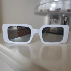 Vita solglasögon från Burberry - Snygga vita solglasögon från Burberry med rektangulära linser och en stilren design. Glasögonen har en elegant gulddetalj med Burberry-loggan på skalmarna. Perfekta för att ge en lyxig touch till din stil.