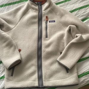 Beige fleecejacka från Patagonia - Säljer en mysig beige fleecejacka från Patagonia med grå dragkedjor och detaljer. Jackan har en hög krage och tre fickor med dragkedja, perfekt för kyliga dagar. Den är långärmad och har en stilren design.