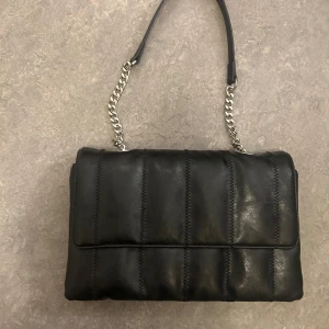 Svart axelväska med kedjedetalj - Snygg svart axelväska i skinn med kedjedetaljer på axelremmen. Väskan har en quiltad design och stängs med tryckknappar. Perfekt för att bära dina nödvändigheter med stil.