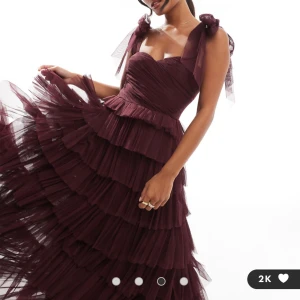 Vinröd långklänning med volanger - Elegant vinröd långklänning med flera lager av volanger och breda axelband med rosetter. Perfekt för en glamorös kväll. Klänningen har en vacker struktur och en feminin siluett.