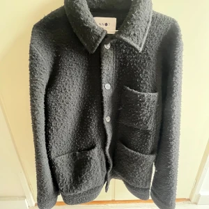 Svart jacka från NN07 - Snygg svart jacka från NN07 med en texturerad yta och knappar framtill. Jackan har en klassisk krage och flera praktiska fickor. Perfekt för en stilren look. Finns ingen storlek på men passar mig bra som M