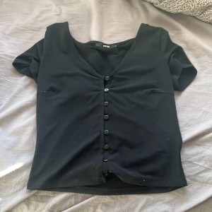 Svart kortärmad topp från Bik Bok - Säljer en svart kortärmad topp från Bik Bok med knappar framtill och v-ringning. Perfekt för en stilren look.