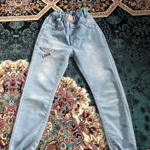 Ljusblå jeansbyxor med broderad detalj - Säljer ett par ljusblå jeansbyxor med resår i midjan och broderad patch med texten 'Love' på ena benet. Byxorna har fickor både fram och bak samt lätt slitna detaljer för en cool look. För barn som är 8 år gammal.