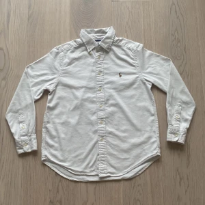 vit ralph lauren skjorta🤍 -  🌟 Vit Stilig Skjorta från Ralph Lauren 🌟  Säljer en elegant vit skjorta från Ralph Lauren, perfekt för alla tillfällen! Den har bara använts ett fåtal gånger och är i mycket bra skick, utan några tydliga defekter. ✨  👕 Specifikationer: - Material: 50% bomull, 50% nylon - Storlek: M (barn)👧🏼🧒🏼 - Modell: Performance   Denna skjorta kombinerar stil och komfort, vilket gör den till ett utmärkt val för både festliga och vardagliga tillfällen😁