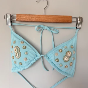 Ljusblå bikiniöverdel med pärldetaljer - Supersöt ljusblå bikiniöverdel med knyt i nacken och ryggen. Utsmyckad med pärlor, snäckor och broderade detaljer i vitt och guld. Triangelformade kupor och trendig design som sticker ut på stranden.