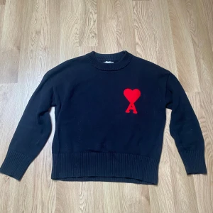 Svart stickad tröja med hjärtlogga - Svart stickad tröja med rund halsringning och ett stort rött hjärta med bokstaven A broderat på bröstet. Tröjan har långa ärmar och ribbade muddar vid ärmslut och nederkant. Perfekt för en stilren och trendig look.