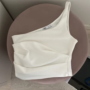 Vit oneshoulder topp från Zara - Säljer en stilren vit oneshoulder topp från Zara. Toppen har en asymmetrisk design med en axel och är tillverkad i ett mjukt, stretchigt material som sitter snyggt på kroppen. Perfekt för att skapa en trendig look.