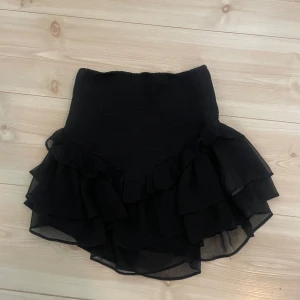 Svart volangkjol från Gina  - Supersöt svart kjol med flera lager volanger och elastisk midja. Kjolen är kort och har en luftig känsla tack vare det tunna materialet. Perfekt att styla med en enkel topp eller stickad tröja.