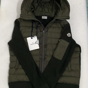 moncler cardigan - Snygg grön pufferjacka från Moncler med stickade ärmar och huva. Jackan har dragkedja framtill, ribbade muddar och en liten ficka på ena ärmen. Perfekt för kyliga dagar när du vill vara både varm och stilren. NFC tag funkar