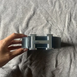 Svart skärp med silverfärgat H-spänne - Säljer ett svart bälte i skinn med ett stort, silverfärgat H-format spänne. Bältet har en stilren och klassisk design med präglad yta och passar perfekt till jeans eller kostymbyxor.Har gjort ett eget hål med verktyget som följer med, annars helt nytt.
