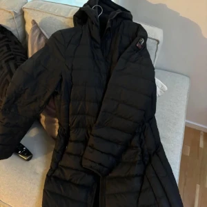 Svart lång dunjacka från Parajumpers - Säljer en svart lång pufferjacka från Parajumpers med huva och dragkedja. Jackan har en klassisk quiltad design och logotyp på ärmen. Perfekt för kyliga dagar.