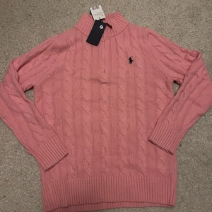 Rosa stickad polotröja från Ralph Lauren - Säljer en rosa stickad polotröja från Ralph Lauren med kabelstickat mönster och halv dragkedja vid halsen. Tröjan har långa ärmar och den klassiska loggan broderad på bröstet. Perfekt för en snygg och avslappnad stil.