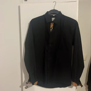 Svart skjorta från Burberry med rutiga detaljer - Säljer en svart skjorta från Burberry med klassiska beige-rutiga detaljer. Säljer för att den inte kommer till användning längre. Den är i storlek XL men passar mest L.