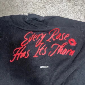 Svart skjorta med tryck från Represent - Cool svart skjorta från Represent med stort grafiskt tryck på ryggen och texten 'Every Rose Has Its Thorn' i rött. Framsidan har en färgstark illustration och logga. Skjortan har klassisk krage och är tillverkad i bomull.