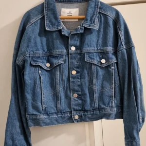 Blå jeansjacka från H&M - Klassisk blå jeansjacka med silverfärgade knappar och två bröstfickor med lock. Jackan har krage och raka sömmar, perfekt för en avslappnad stil. Croppad modell som ger en modern look.