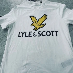 Vit t-shirt från Lyle & Scott - Säljer en fräsch vit t-shirt från Lyle & Scott med stor gul örn och svart logga på bröstet. Klassisk passform och rund halsringning. Perfekt till jeans eller shorts!