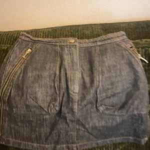 Mörkblå jeanskjol med dragkedjor - Säljer en mörkblå jeanskjol med flera dragkedjor både fram och bak. Kjolen har en klassisk jeanslook med knapp i midjan och dekorativa sömmar. Perfekt för dig som gillar en edgy stil.