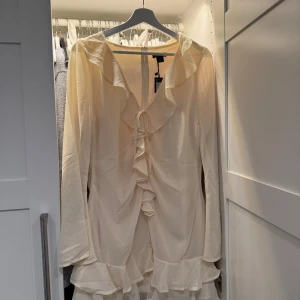 Krämvit volangklänning med v-ringning - Säljer en krämvit klänning med långa ärmar och v-ringning. Klänningen har volangdetaljer längs halsringningen och nederkanten samt en knytning framtill. Materialet känns lätt och flowy, perfekt för sommaren. Nypris 399kr säljer nu för 225