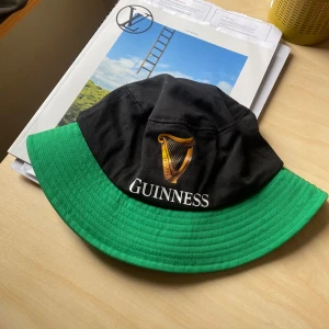 Svart och grön bucket hat från Guinness - Bucket hat i svart och grönt med Guinness-logga och harpa broderad framtill. Klassisk form med bred, nedvikt brätte. Perfekt accessoar för dig som gillar ikoniska märken och vill sticka ut. ONE SIZE