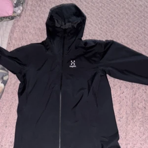 Haglöfs Svart vindjacka  - Svart vindjacka från Haglöfs med huva och dragkedja framtill. Jackan har lång ärm och är tillverkad i syntetmaterial. Perfekt för utomhusaktiviteter och skyddar mot vind och lätt regn. (HELT OANVÄND)