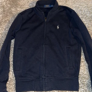 tröja från Polo Ralph Lauren - Säljer en svart tröja från Polo Ralph Lauren med dragkedja framtill och ståkrage. Jackan har två fickor på sidorna och den klassiska loggan broderad på bröstet. Perfekt för en stilren look.
