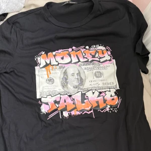 Svart t-shirt med tryck 'Money Talks' - Cool svart t-shirt med stort tryck av en dollarsedel och texten 'Money Talks' i graffiti-stil med rosa och orange detaljer. Perfekt statement-plagg för dig som gillar streetwear och unika prints.