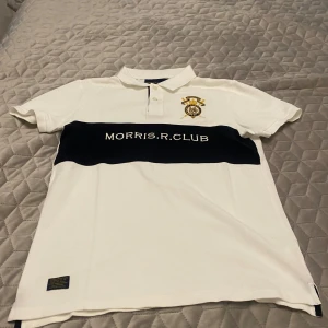 Vit piké från Morris R. Club - Vit pikétröja från Morris R. Club med klassisk krage och knappar. Stor broderad logga i gult och blått på bröstet samt en bred marinblå rand med texten 'MORRIS.R.CLUB' över bröstet. Perfekt för en stilren och sportig look.