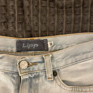 Ljusblå jeansshorts från Lipp - Säljer ett par ljusblå jeansshorts från Lipp med råa kanter och klassisk femficksdesign. Shortsen har hög midja och en avslappnad passform, perfekta för varma dagar. Stängs med knapp och dragkedja framtill.