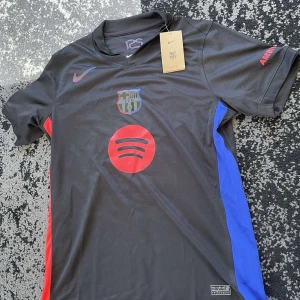 FC Barcelona svart bortatröja Nike - Säljer en svart FC Barcelona bortatröja från Nike med röda och blå detaljer på sidorna. Tröjan har klubbmärke och Spotify-logga på bröstet samt Nike-logga i rött. Kortärmad modell i lätt och ventilerande material.