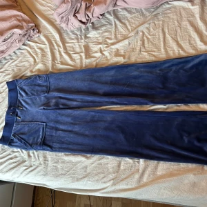 Blå mjukisbyxor från Juicy Couture - Säljer ett par blå mjukisbyxor från Juicy Couture med bred resår i midjan och snörning. Byxorna har två fickor med lock och knappar bak samt en broderad Juicy-logga på ena fickan fram. Perfekta för en avslappnad stil. Bara använda ett fåtal gånger💗
