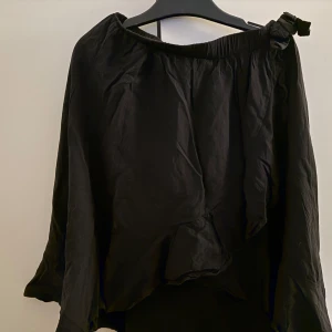 Svart offshoulder blus med rosett - Säljer en svart offshoulder blus med en dekorativ rosett på ena axeln. Blusen har en lös passform och är tillverkad i ett lätt, mjukt tyg som faller fint. Perfekt för dig som gillar stilrena och trendiga plagg.