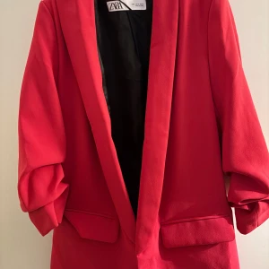 Röd kavaj från ZARA - Säljer en snygg röd kavaj från ZARA i storlek S. Kavajen har lång ärm, två fickor framtill och en klassisk krage. Perfekt för att lyfta vilken outfit som helst med en pop av färg.