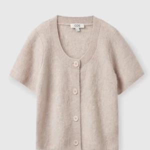 Beige kortärmad kofta från COS - Superfin beige kofta från COS med korta ärmar och stora knappar framtill. Tröjan är stickad och har en croppad passform som passar perfekt till jeans eller kjol. Enkel och stilren design som funkar till många outfits.
