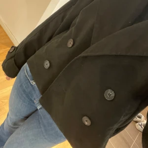 Trenchcoat  - Säljer en svart trenchcoat från H&M med inga defekter. Jackan har långa ärmar och stora knappar framtill. Passar perfekt till våren och sommaren!