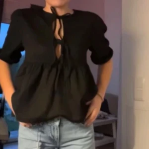 Svart volang blus med knytning  - Säljer en svart kortärmad blus med knytning framtill. Blusen har volang nedtill och är tillverkad i ett ribbat material. Aldrig använd. 