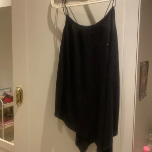 Svart asymmetrisk topp från Anbar - Säljer en svart klänning från Anbar med tunna axelband och asymmetrisk nederkant. Klänningen har en enkel och stilren design, perfekt för dig som gillar minimalistiskt mode. Materialet känns mjukt och följsamt.