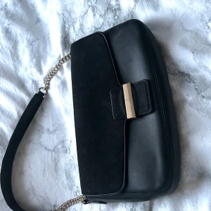Svart axelväska med gulddetalj - Snygg svart axelväska med kedjerem och guldfärgat spänne framtill. Väskan har en stilren design med både släta och mockaliknande partier. Perfekt för att lyfta din outfit med en elegant touch.