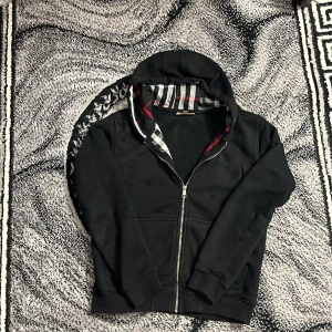 Svart hoodie med dragkedja från Burberry - Säljer en svart hoodie från Burberry med dragkedja och klassiskt rutigt foder i huvan. Tröjan har långa ärmar, fickor framtill och en diskret broderad logga på bröstet. Perfekt för en stilren och bekväm look.