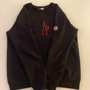 Svart sweatshirt från Moncler - Svart sweatshirt från Moncler med broderad logga och röd detalj framtill. Klassisk rund halsringning och långa ärmar. Perfekt för en stilren och avslappnad look.