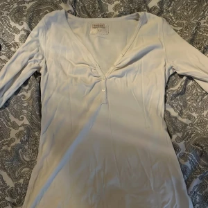 Vit långärmad tröja med knappar från Bik Bok - Säljer en vit långärmad tröja från Bik Bok med v-ringning och dekorativa knappar framtill. Tröjan har en figurnära passform och är tillverkad i ett mjukt, stretchigt material. Perfekt för dig som gillar enkel och stilren design.