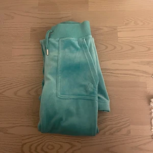 Turkosa mjukisbyxor från Juicy Couture - Säljer ett par turkosa mjukisbyxor från Juicy Couture med broderad logga på fickan och dragsko i midjan. Byxorna är i mjukt material och har bred mudd upptill. Perfekta för en avslappnad stil.