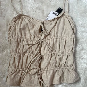 Beige linnetopp med knytning från Divided - Supersöt beige linnetopp från Divided i linneblandning. Toppen har smala axelband, volangkant nertill och snygga knytningar framtill. Perfekt för varma dagar och enkel att styla med jeans eller kjol.