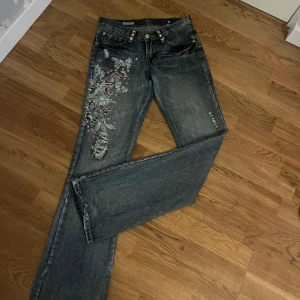 Broderade jeans från 7 For All Mankind x Zac Posen - Säljer ett par lågmidjade ljusblå boutcut jeans med broderade blommor och slitningar på ena benet. Någon slitning längs ner! 