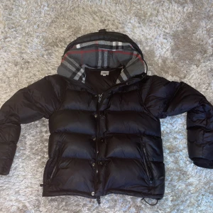 Svart dunväst/jacka från Burberry med avtagbara armar. - Säljer en svart dunväst från Burberry i storlek S med klassiskt rutigt foder i huvan. Västen har dragkedja och knappar framtill samt två fickor med dragkedja. Jackan saknar 1 knapp men kan enkelt sättas på en ny. Kontakta för mer bilder osv.