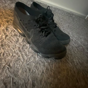 Svarta Nike Vapormax sneakers - Säljer ett par helsvarta Nike Vapormax sneakers med stickad ovandel och snörning. Skorna har den ikoniska bubbliga sulan och en sportig look som passar perfekt till vardags eller träning.