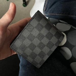 Rutig plånbok från Louis Vuitton - Snygg plånbok från Louis Vuitton i klassiskt rutigt mönster. Plånboken är svart och grå, tillverkad i skinn med flera kortfack och plats för ID-kort. Perfekt accessoar för dig som vill ha stilren design. Kvitto finns!