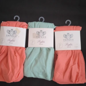 Topeco tights i korall och mint - Tre par Topeco tights i one size, två i korallrosa och ett i mintgrönt. Tightsen är tunna, 60 denier, och tillverkade i 90% polyamid och 10% elastan för en mjuk och stretchig känsla. Perfekta för att addera färg till din outfit.