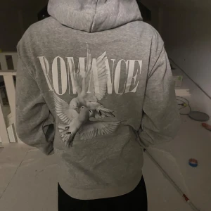 Grå hoodie med duvor och text - Säljer en grå hoodie med stor vit text 'ROMANCE' och två detaljerade duvor tryckta på ryggen. Tröjan har huva och en avslappnad passform, perfekt för dig som gillar streetwear vibes och snygga prints.