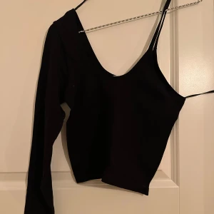 Svart asymmetrisk topp från STR - Säljer en svart, riktigt cool asymmetrisk topp från STR. Ena sidan har lång ärm och andra sidan är ärmlös med smal axelrem, vilket ger en unik och trendig look. Perfekt för dig som vill sticka ut med en edgy stil.